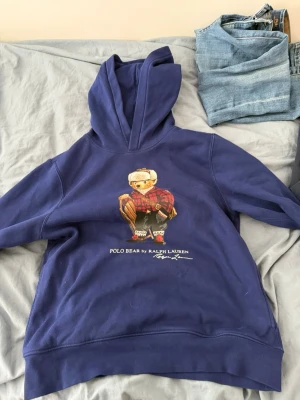 Blå hoodie Polo Bear Ralph Lauren - Säljer en blå hoodie från Ralph Lauren med den ikoniska Polo Bear-trycket på bröstet. Hoodien har huva och ribbade muddar. Tillverkad i mjukt bomullsmaterial och har en avslappnad passform. Perfekt för en chill och stilren look.