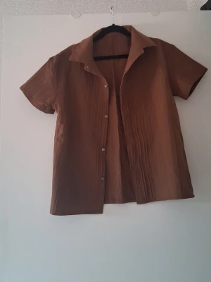 Brun kortärmad skjorta med knappar och shorts - Säljer en brun skjorta med korta ärmar och knappar framtill. Skjortan har en avslappnad passform och är tillverkad i ett lätt, strukturerat tyg som ger en snygg look. Perfekt till sommaren och enkel att styla med olika outfits.
