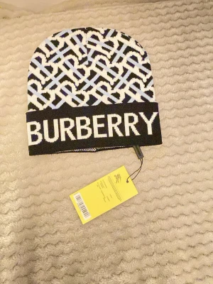 Burbǝrry svartvit  mössa - Säljer en snygg svartvit mössa från Burbǝrry med stort logotyp-tryck framtill och ett grafiskt mönster i svart, vitt och ljusblått. Mössan är ribbstickad och har en uppvikt kant. Perfekt accessoar för att lyfta din outfit.