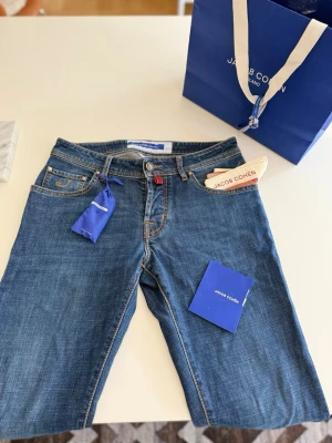 Blå jeans från Jacob Cohën - (ALDRIG ANVÄNDA, KVITTO FINNS) KÖPTA  i PARIS.       Snygga blå jeans från Jacob Cohën med klassisk femficksmodell och kontrastsömmar. Jeansen har en liten broderad J på myntfickan och detaljer i silver. Tillverkade i mjukt denimtyg för en bekväm passform. Perfekta för en avslappnad och trendig stil.  MODELL: NICK SLIM 31. 