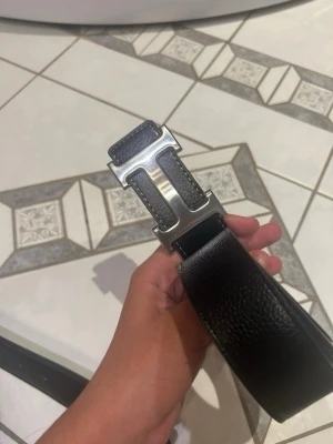 Svart skärp med silverfärgat H-spänne - Snyggt svart bälte i skinn med ett stort, silverfärgat H-format spänne. Perfekt accessoar för att lyfta vilken outfit som helst och ger en clean och stilren look.