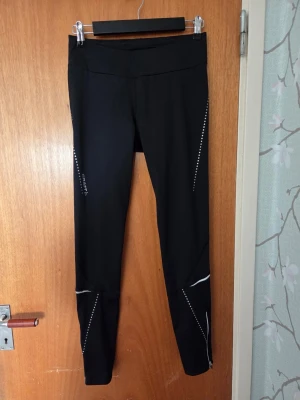 Svarta sportleggings med reflexdetaljer - Säljer ett par svarta sportleggings med vita och reflekterande detaljer längs benen. De har hög midja och är tillverkade i ett stretchigt material som passar perfekt för träning eller löpning. Snygg och slimmad passform med diskreta mönster på sidorna.