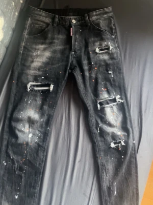 Svarta slitna jeans med färgstänk - Säljer ett par svarta jeans med slitningar och hål på benen samt vita och orange färgstänk för en edgy look. Passar också size 48 som står för W30 fast egentligen är de W29. Jeansen har klassisk femficksmodell och raka ben. Perfekta för dig som gillar streetwear och vill sticka ut. 