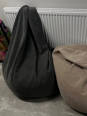 Två stora sittsäckar i tyg - Säljer två stora sittsäckar, en i mörkgrått tyg och en i beige tyg. Perfekta för att chilla i vardagsrummet eller spelrummet. De är mjuka och formar sig efter kroppen för maxad komfort. Passar både till filmkvällar och gaming.