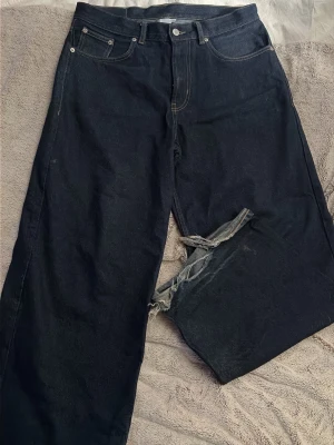 Mörkblå pösiga raka junkyard jeans med slitningar - Säljer ett par mörkblå jeans med raka ben och tydliga slitningar på ben änden. Jeansen har klassiska kontrastsömmar. Perfekta för dig som gillar en edgy och avslappnad stil.