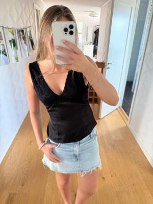 Svart linnetopp med spetsdetaljer - Snygg svart linnetopp med v-ringning och spetsdetaljer upptill. Toppen är ärmlös och har en figurnära passform som ger en stilren och trendig look. Perfekt att matcha med jeans eller kjol för en cool outfit.
