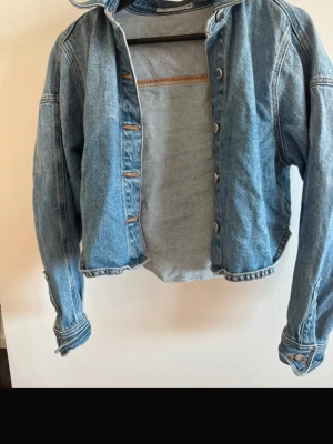 Cropped blå jeansjacka - Snygg cropped jeansjacka i ljusblå denim med klassisk krage och knappstängning framtill. Jackan har långa ärmar med knappar vid ärmsluten och en avslappnad passform. Perfekt för dig som vill ha en trendig och avslappnad look.