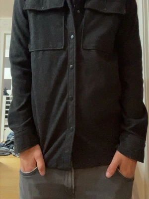 Mörkgrå skjorta/overshirt med bröstfickor - Stilren mörkgrå skjorta med lång ärm och två stora bröstfickor med lock och knapp. Skjortan har klassisk krage och knäppning framtill. Materialet ser ut att vara mjukt och lite tjockare, perfekt för lager-på-lager. Passar dig som gillar en enkel men snygg look.