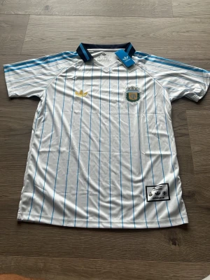 Argentina fotbollströja Adidas - Snygg Argentina fotbollströja från Adidas i vitt med tunna blå ränder. Tröjan har korta ärmar, blå detaljer vid kragen och axlarna samt broderad landslagssköld och Adidas-logga. Tillverkad i 100% polyester, perfekt för träning eller match.