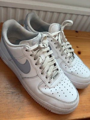 Nike Air Force 1 vita sneakers -  Klassiska Nike Air Force 1 sneakers i vitt läder med ljusgrå detaljer och perforerad tå. Ikonisk Swoosh-logga på sidan, AIR-detalj på hälen och vita snören.  Clean och stilren modell som passar till det mesta 🤍  Skorna är använda och har normalt slitage, se bilder för exakt skick.