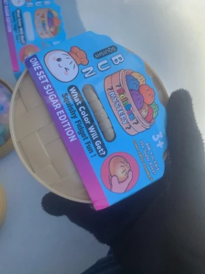 Squishy Bun Mystery Sugar Edition - Squishy-leksak i form av en bun, levereras i en rund beige plastförpackning med färgglad etikett. Innehåller en överraskningsfärgad squishy som är mjuk och skön att klämma på. Perfekt som fidget toy eller stressleksak. För barn från 3 år och uppåt.
