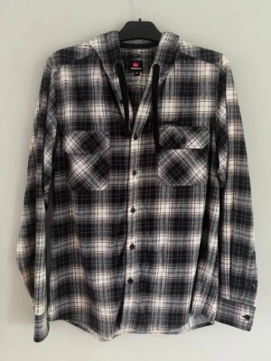 Quiksilver rutig overshirt/jackskjorta med luva - Overshirt från Quiksilver i storlek M. Svart, brungrått och vitt rutigt mönster med knappstängning och två bröstfickor. Har en luva med mjukt grått tyg på insidan och svarta snören. Tillverkad i 100% bomull och känns väldigt mjuk. Mid fit passform. Mycket bra skick, inga skador eller annat.