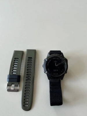 Garmin Fenix 6 PRO - Säljer en Garmin Fenix smartklocka i svart med robust boett och tydlig display. Klockan har utbytbara armband, ett i grönt silikon och ett i svart tyg. Perfekt för dig som gillar träning och äventyr, med funktioner som höjdmätare och stegräknare.