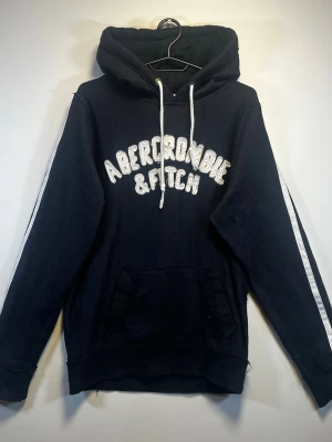 Abercrombie & Fitch Vintage Muscle Hoodie – Navy - Klassisk Abercrombie & Fitch hoodie i mörk marinblå färg med stort broderat/applikationslogo framtill och vita ränder längs ärmarna. Äldre Muscle-modell med riktigt snygg vintage/Y2K-look.  Storlek: L Färg: Mörkblå / Navy Material: 100% bomull  Välanvänt vintage-skick med tydligt slitage på bland annat ficka, sömmar och muddar – se bilder. Slitaget ger hoodien en naturligt distressed/vintage look.