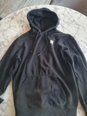 Svart hoodie med vit patch - Svart hoodie med stor vit fyrkantig patch på ryggen och en mindre vit patch på bröstet. Klassisk känguruficka framtill och justerbar huva med dragsko. Perfekt för dig som gillar enkel men unik streetwear-stil.