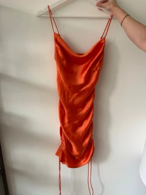 Orange klänning med snörning - Snygg orange kortklänning i satinliknande material med tunna axelband och draperad framsida. Klänningen har snörningsdetaljer på sidan. Oanvänd, prislapp kvar. 