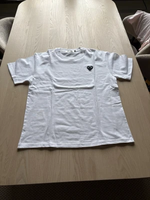 Vit Comme des Garçons PLAY t-shirt - Säljer en vit t-shirt från Comme des Garçons PLAY med det ikoniska svarta hjärtat med ögon broderat på bröstet. T-shirten har rund hals och korta ärmar. Tillverkad i mjuk bomull som känns skön mot huden.