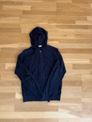 Mörkblå hoodie med dragkedja från RYVLS - En stilren mörkblå hoodie med dragkedja framtill och huva. Hoodien har långa ärmar och ribbade muddar vid ärmslut och nederkant. Perfekt för en avslappnad look och enkel att matcha med andra plagg.