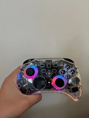 Transparent LED-handkontroll Deltaco - Säljer en transparent handkontroll från Deltaco Gaming med LED-belysning i regnbågsfärger runt styrspakarna. Kontrollern har tydliga knappar, två styrspakar och D-pad. Tillverkad i plast med synliga kretskort och svart detaljering. Perfekt för gaming med cool look. Fixade gammalt stick drift som inte är kvar😁kommer med en usb c kontakt så man kan plugga in den