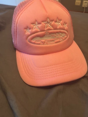 Rosa meshkeps med broderi - Snygg rosa keps med mesh på baksidan och justerbar snapback. Framtill finns broderade stjärnor och ett motiv, samt broderad text på sidan. Perfekt för en trendig och avslappnad stil.