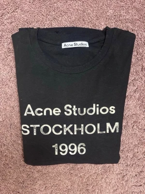 Svart långärmad t-shirt Acne Studios Stl S - Snygg svart långärmad t-shirt från Acne Studios med tryck 'Acne Studios STOCKHOLM 1996' i vitt framtill. Klassisk rund hals och rak passform. Perfekt för dig som gillar stilrena och trendiga plagg med streetkänsla.