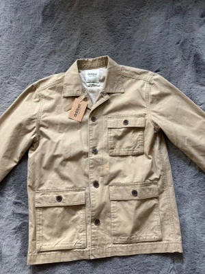 Beige overshirt från Dobber - Stilren beige overshirt från Dobber med tre stora fickor framtill och knappar i mörkbrunt. Klassisk krage och rak passform i ett slitstarkt bomullstyg. Perfekt lager-på-lager-plagg för en avslappnad look. Storlek S passar M