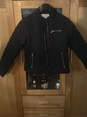 Svart quiltad jacka från SAMSA Collection - Snygg svart quiltad jacka från SAMSA Collection med dragkedjor framtill och på fickorna. Jackan har en modern, kortare passform och långärmade ärmar. Perfekt för dig som vill ha en stilren och trendig look.