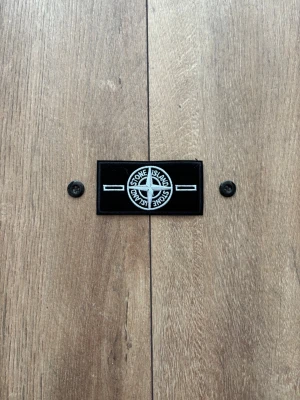 Stone Island Patch / Badge – Svart med vit/silver brodyr & knappar - Säljer en klassisk Stone Island-badge i svart med vit/silverfärgad brodyr, inklusive tillhörande originalknappar precis som på bilderna. Patchen är helt ny, oanvänd och i perfekt skick. Passar utmärkt som reservdel eller tillbehör till din Stone Island-jacka eller tröja. 