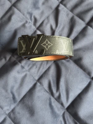 Svart Louis Vuitton monogram skärp - Säljer ett svart skärp från Louis Vuitton med klassiskt monogrammönster och stor LV-logga i metall som spänne. Bältet har en stilren design och är tillverkat i skinn med en snygg struktur. Perfekt accessoar för att lyfta din outfit.