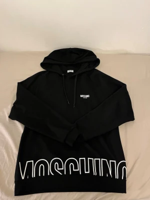 Svart hoodie från Moschino - Snygg svart hoodie från Moschino med huva och dragsko. På bröstet står det 'Moschino Active' i vitt och längs nederkanten finns ett stort vitt Moschino-tryck. Hoodien har en avslappnad passform och är tillverkad i mjukt material.