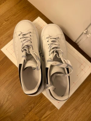 Vita chunky sneakers med svart detalj - Snygga vita sneakers med chunky sula och svart detalj på hälen. Skorna har vita snören med svart mönster och perforerade sidor för extra stil. Tillverkade i slätt material som ger en clean look och passar perfekt till streetwear.