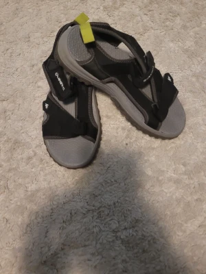 Svarta och gråa sandaler från Quechua - Säljer ett par sportiga sandaler från Quechua i svart och grått med justerbara kardborreband och en cool gul detalj på hälen. Perfekta för äventyr och utomhusaktiviteter. Sulan är robust och ger bra grepp.