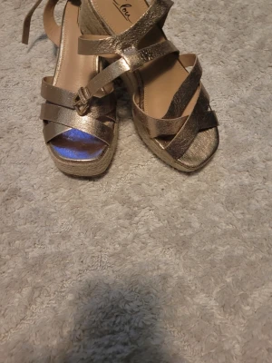 Guldiga platåsandaler med remmar - Säljer ett par snygga sandaler i metallic guld med flera korsade remmar och justerbart spänne. Sandalerna har platåsula i flätad look och öppen tå. Perfekta för dig som vill sticka ut lite extra med din outfit.