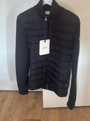 Svart Cardigan dunjacka från Moncler - Snygg svart dunjacka från Moncler med quiltad front, dragkedja och två fickor med dragkedja. Jackan har Moncler-logga på ärmen och ribbade muddar. Perfekt för kalla vinterdagar och riktigt trendig stil. Storlek M, aldrig använd bara testad och är för stor för mig