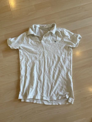 Vit piké t-shirt  - Vit piké t-shirt med klassisk krage och korta ärmar. Tröjan har knappar vid halsen och en liten broderad detalj på bröstet. Tillverkad i mjuk bomull, perfekt enkel stil.