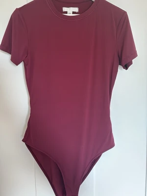 Vinröd body från H&M, kort ärm - Säljer en vinröd body från H&M i storlek XS. Bodyn har korta ärmar, rund halsringning och är gjord i ett mjukt, stretchigt material som sitter snyggt på kroppen. Perfekt att styla med jeans eller kjol för en clean look.