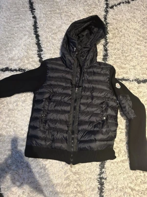 Svart dunväst med huva från Moncler - Snygg svart dunväst från Moncler med huva och stickade ärmar. Västen har två fickor med knappar framtill och dragkedja. Klassisk Moncler-logga på ena ärmen. Perfekt för lager på lager och streetstyle. Priset är inte fast och cardiganen passar 170cm 