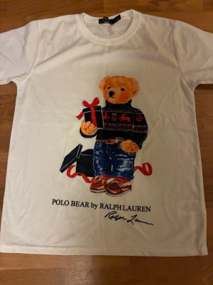 Vit Polo Bear t-shirt från Ralph Lauren - Vit t-shirt från Ralph Lauren med ikoniskt Polo Bear-motiv på framsidan. Björnen har på sig en stickad tröja, jeans och håller i ett presentpaket. T-shirten har rund hals och korta ärmar. Perfekt för dig som gillar klassiska och lekfulla prints.            Kontakta gärna vid frågor och prisförslag. Skickar inom 24h. 