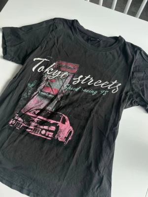T-shirt med tryck från Gina Tricot - En mörkgrå t-shirts med ”Tokyo Streets” racing-tryck. Storlek S. Mycket bra skick!
