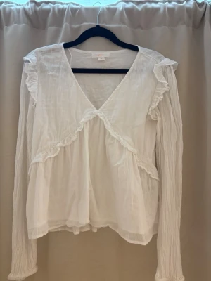 Vit volangblus från Gina Tricot - Söt vit blus från Gina Tricot med djup V-ringning, volangdetaljer framtill och långa, lätt transparenta ärmar. Tillverkad i ett tunt, luftigt tyg som ger en romantisk känsla. Perfekt för dig som gillar en feminin och trendig stil.