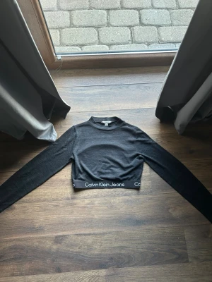 Svart croppad topp Calvin Klein - Snygg svart croppad topp från Calvin Klein Jeans med lång ärm och elastisk kant med logga nedtill. Perfekt till höga jeans eller kjol. Enkel och stilren design med en tight passform.