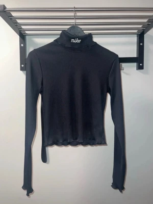 Svart ribbad polotopp från Nike - Säljer en svart ribbad polotopp från Nike med broderad logga på kragen. Toppen har lång ärm och croppad passform med vågig kant nertill. Perfekt för dig som gillar sportig och stilren look. Den är aldrig använd.