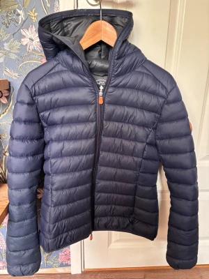 Mörkblå dunjacka med huva - Snygg mörkblå dunjacka från Save The Duck med huva och dragkedja framtill. Jackan har quiltad design, smal passform och en rund orange logotyp på ärmen. Perfekt för kalla vinterdagar och riktigt lätt att bära.