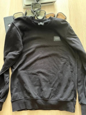 Svart sweatshirt från Dolce & Gabbana - Stilren svart sweatshirt från Dolce & Gabbana med en metallbricka på bröstet där loggan står. Tröjan har rund halsringning, långa ärmar och ribbade muddar vid ärmslut och nederkant. Perfekt för en clean och trendig look.