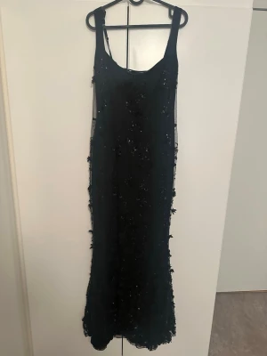 Svart långklänning med snörning - Svart långklänning med tunna axelband, dekorerad med paljetter och blommiga detaljer. Klänningen har snörning i ryggen och är gjord i ett tunt, transparent material med mesh-känsla. Perfekt för dig som vill ha en elegant och unik look.