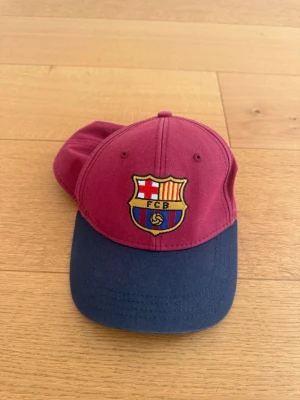 Vinröd och marinblå FC Barcelona keps - Snygg keps i vinrött och marinblått med FC Barcelona-logga broderad framtill och texten 'FC BARCELONA' broderad baktill. Justerbar rem bak för perfekt passform. Tillverkad i bomullstyg, perfekt för dig som älskar fotboll och vill visa ditt lag.