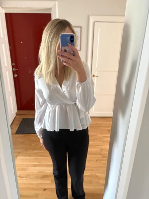 Vit omlottblus med volang - Säljer en vit omlottblus med långa ärmar och volang nedtill. Blusen har en markerad midja och v-ringning framtill. Perfekt för dig som gillar en stilren look. 
