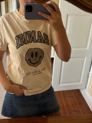 Beige GANNI t-shirt med smiley - Säljer en beige t-shirt från GANNI med svart tryck framtill. T-shirten har ett stort smileyface och texten 'Have a nice day!' samt klassisk rund hals och korta ärmar. Perfekt för en chill och trendig look. Den är i extremt bra skick då bara används fåtal gånger 