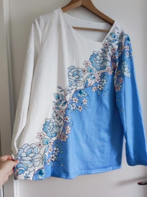 Blommig långärmad topp i blått och vitt - Snygg långärmad topp med asymmetrisk halsringning i vitt och ljusblått. Toppen har ett stort blommigt mönster i blått och rosa som går diagonalt över framsidan. Perfekt för dig som gillar unika detaljer och mjuka färger.