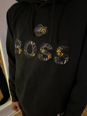 Svart Lakers x BOSS hoodie - Säljer en svart hoodie från BOSS i samarbete med Los Angeles Lakers och NBA. Hoodien har huva med dragsko och tryck med Lakers-logga och texten BOSS i lila, gult och vitt på bröstet. Liten NBA x BOSS-patch på nedre delen. Perfekt för dig som gillar basket och streetwear. Litet hål vid ärmen. Men inget som är märkbart